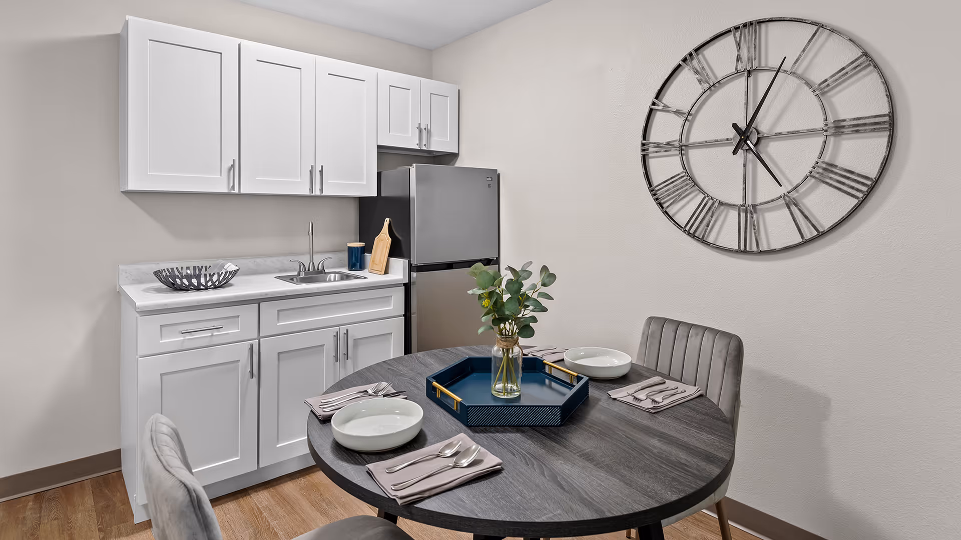 A small kitchen and dining area with white cabinets, a stainless steel refrigerator, a sink, and a round dark wood table set with two place settings, a blue tray, and a vase with green foliage. A large decorative wall clock with Roman numerals is mounted on the wall.