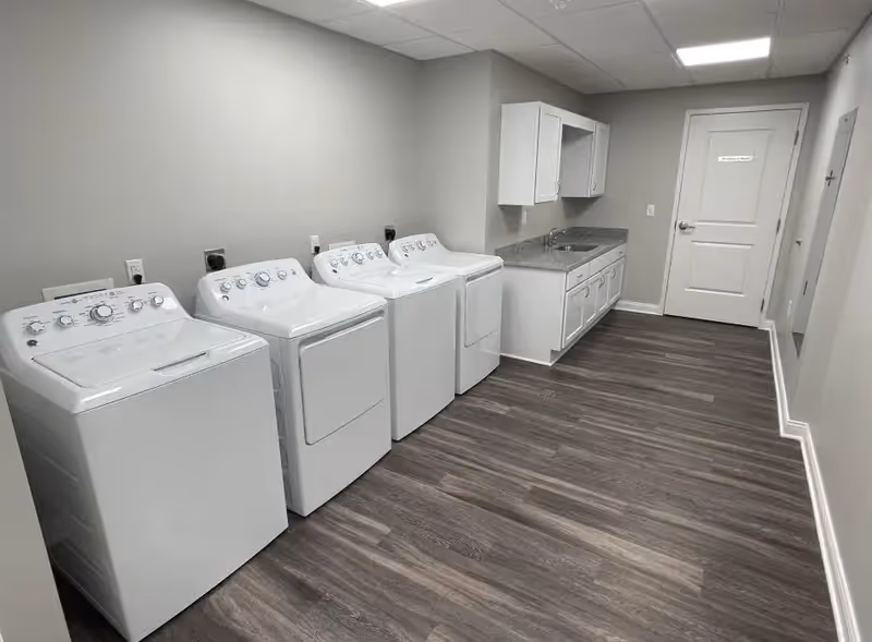 Laundry room with four white washers and dryers lined up along one wall and a sink with cabinets at the far end.