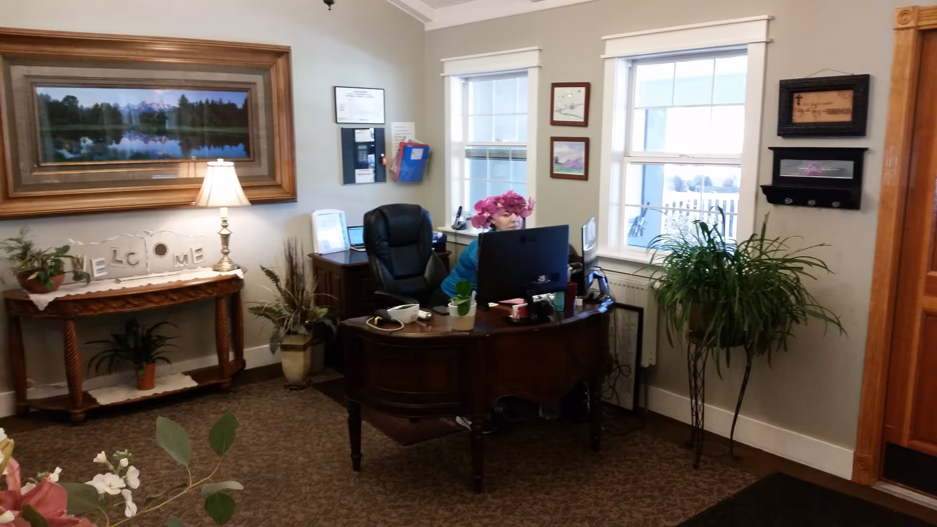 Reception area of The Manor on Marston Lake with a wooden desk, a computer monitor, and a person sitting behind the desk. The room has beige walls, two windows with white trim, several framed pictures on the walls, and various potted plants. A wooden console table with a lamp and a 'WELCOME' sign is against one wall.