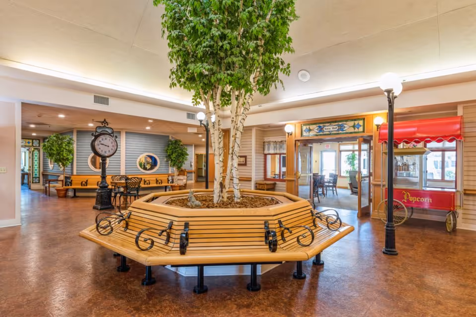Interior view of a senior living facility common area featuring a large octagonal wooden bench surrounding a planter with tall leafy trees. There is a vintage-style black clock on a post, two black streetlamp-style lights, a red popcorn cart, and a doorway leading to a dining hall with tables and chairs. The floor is a warm brown color and the walls have light wood paneling with decorative windows.