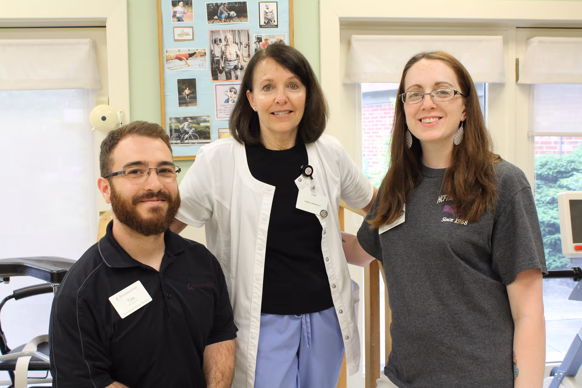 Three staff members standing together inside a room with exercise equipment and a bulletin board with photos on the wall behind them. The man on the left is wearing glasses and a black polo shirt with a name tag that reads 'Tim'. The woman in the middle is wearing a white medical coat over a black shirt and light blue pants. The woman on the right is wearing glasses and a dark gray t-shirt with a logo and text on it. They are all smiling.