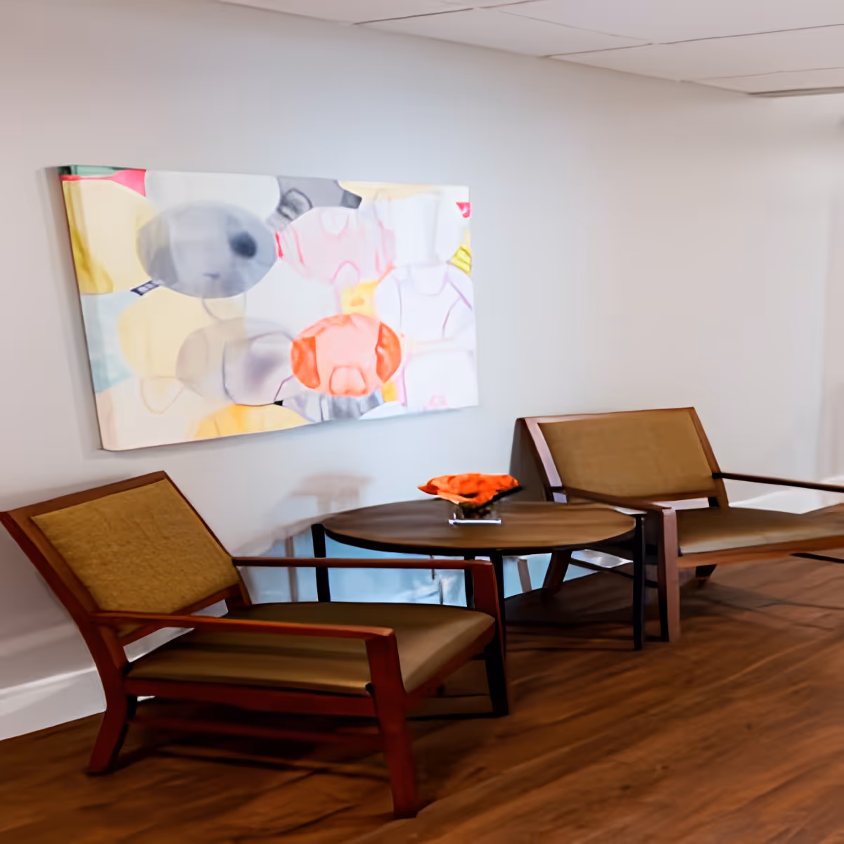 A cozy seating area with two wooden chairs featuring beige cushions and a round wooden table between them. On the table is a decorative orange centerpiece. A colorful abstract painting hangs on the light gray wall behind the chairs. The floor is wooden, and the ceiling has white tiles.