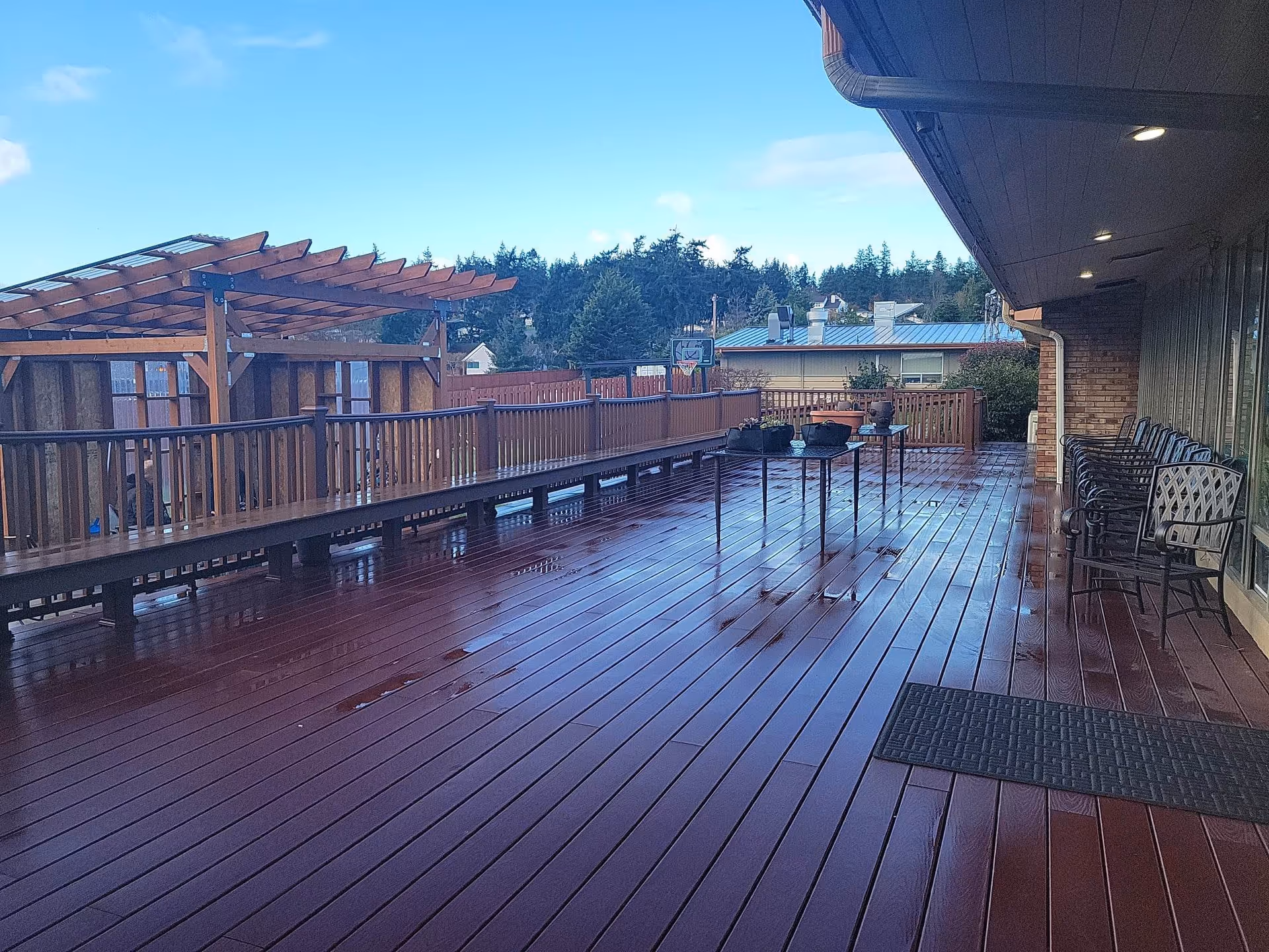 Outdoor wooden deck area with a pergola on the left side, a long bench along the railing, several chairs lined up on the right side near the building, and two small tables with potted plants. Trees and rooftops are visible in the background under a partly cloudy sky.
