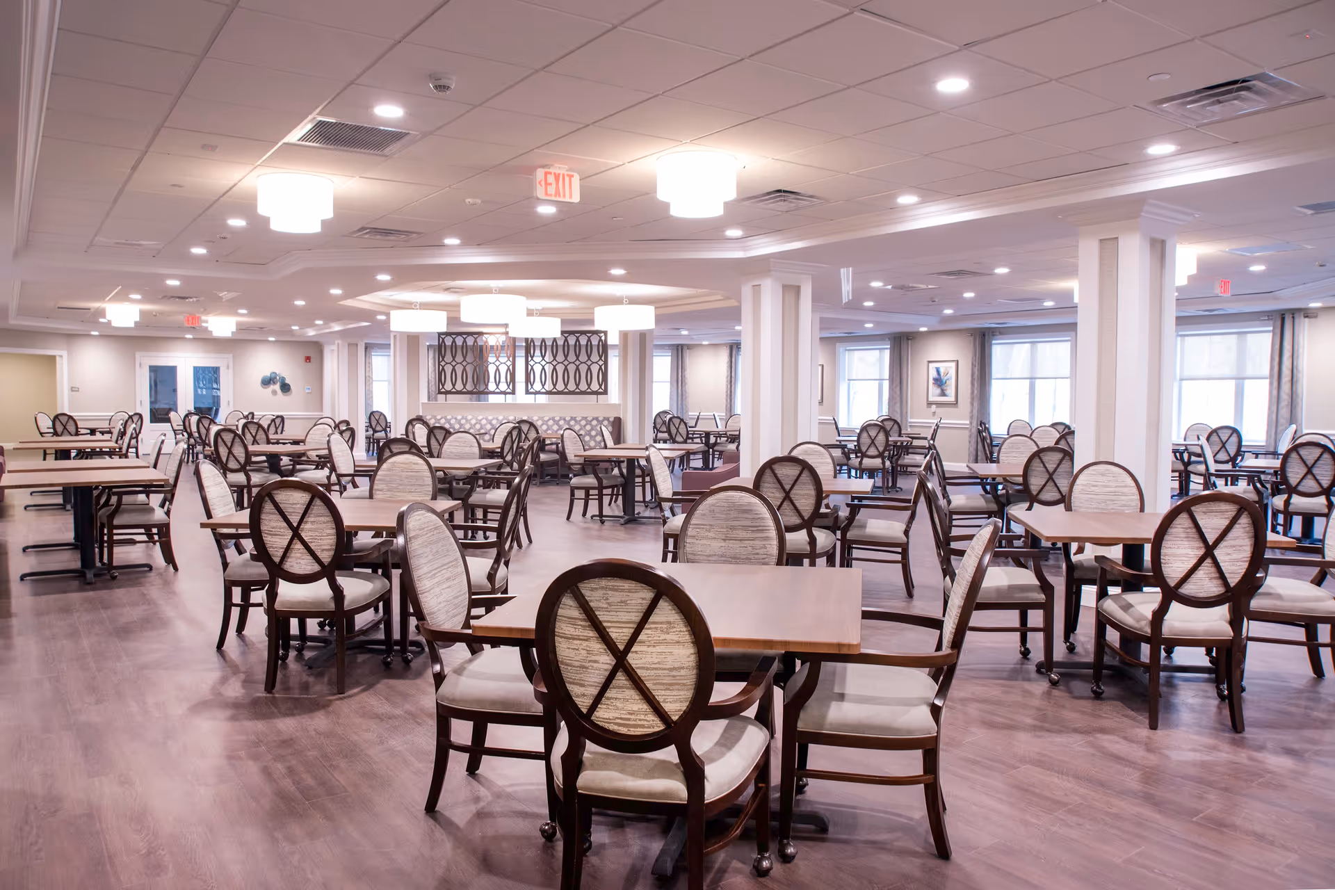 A spacious dining room with multiple wooden tables and cushioned chairs arranged neatly. The room has a polished wooden floor, white ceiling with recessed lighting, and large windows with curtains allowing natural light. Decorative wall art and modern light fixtures add to the elegant ambiance.