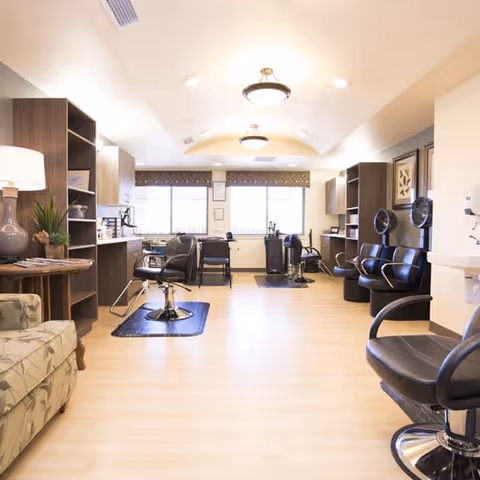 Bright and spacious salon area with multiple black styling chairs, mirrors, and hair drying stations. The room has light wood flooring, beige walls, and large windows with patterned valances letting in natural light. There is a patterned armchair and a side table with a lamp and plant on the left side.