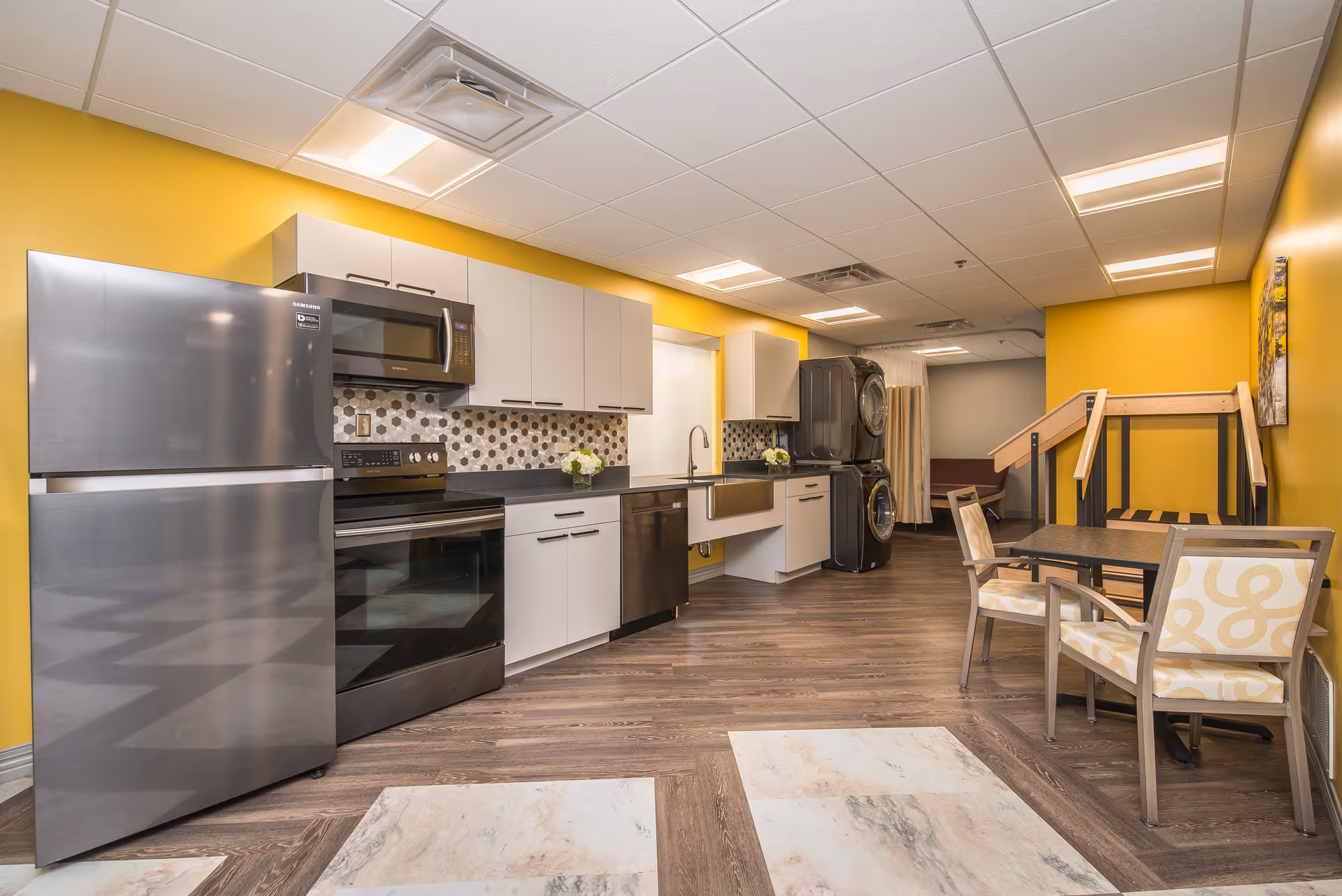 A modern kitchen area with yellow walls, featuring stainless steel appliances including a refrigerator, microwave, stove, and dishwasher. There is a countertop with white cabinets above and below, a farmhouse-style sink, and a stacked washer and dryer in the background. To the right, there is a small dining table with two chairs and a wooden ramp with handrails leading to another room.