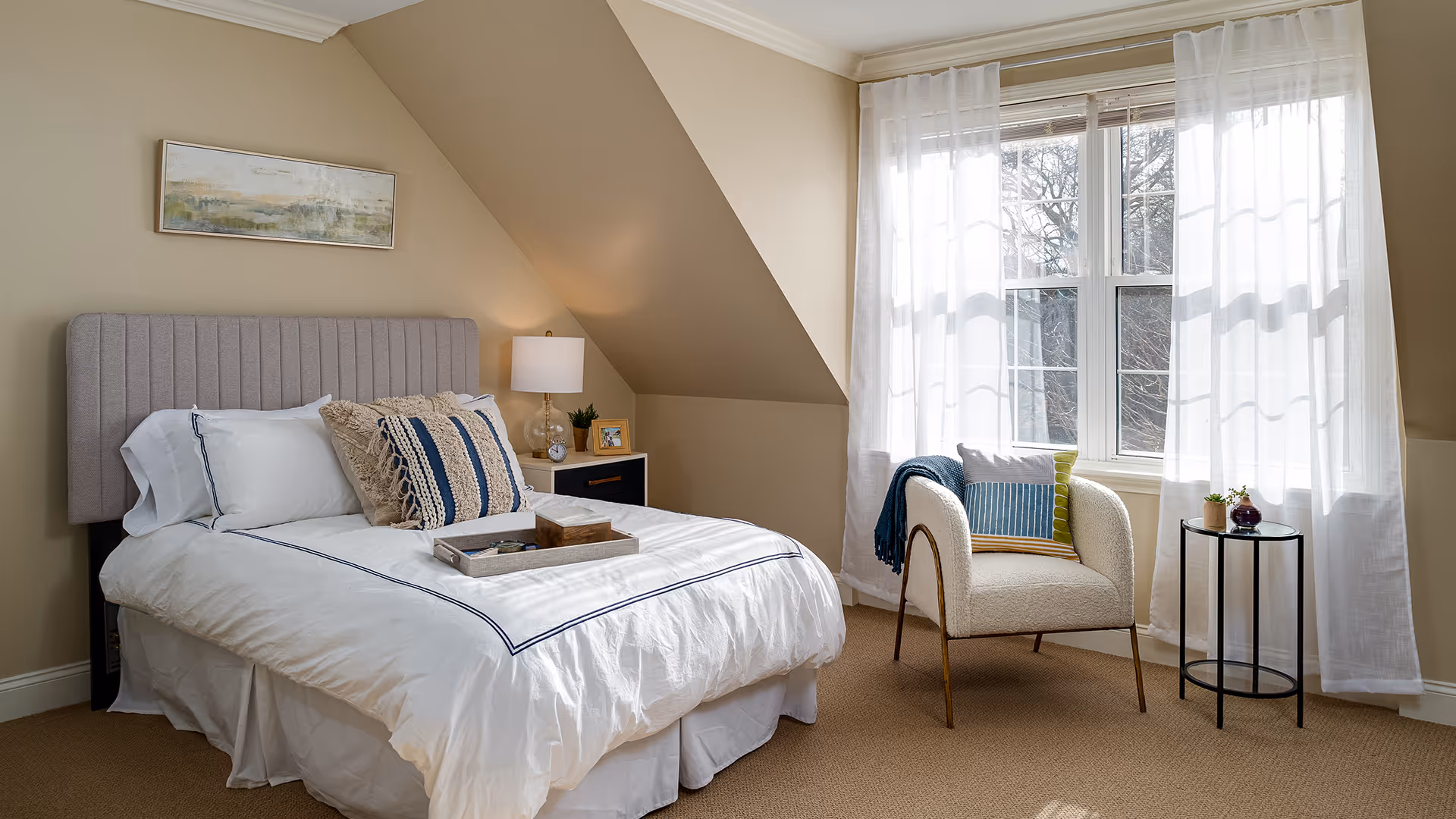 A cozy bedroom with a neatly made bed featuring white bedding and decorative pillows. There is a nightstand with a lamp, clock, and small plant next to the bed. A comfortable armchair with a colorful pillow and a throw blanket is positioned near a window with sheer white curtains, allowing natural light to fill the room. The walls are painted beige, and a framed abstract painting hangs above the bed.