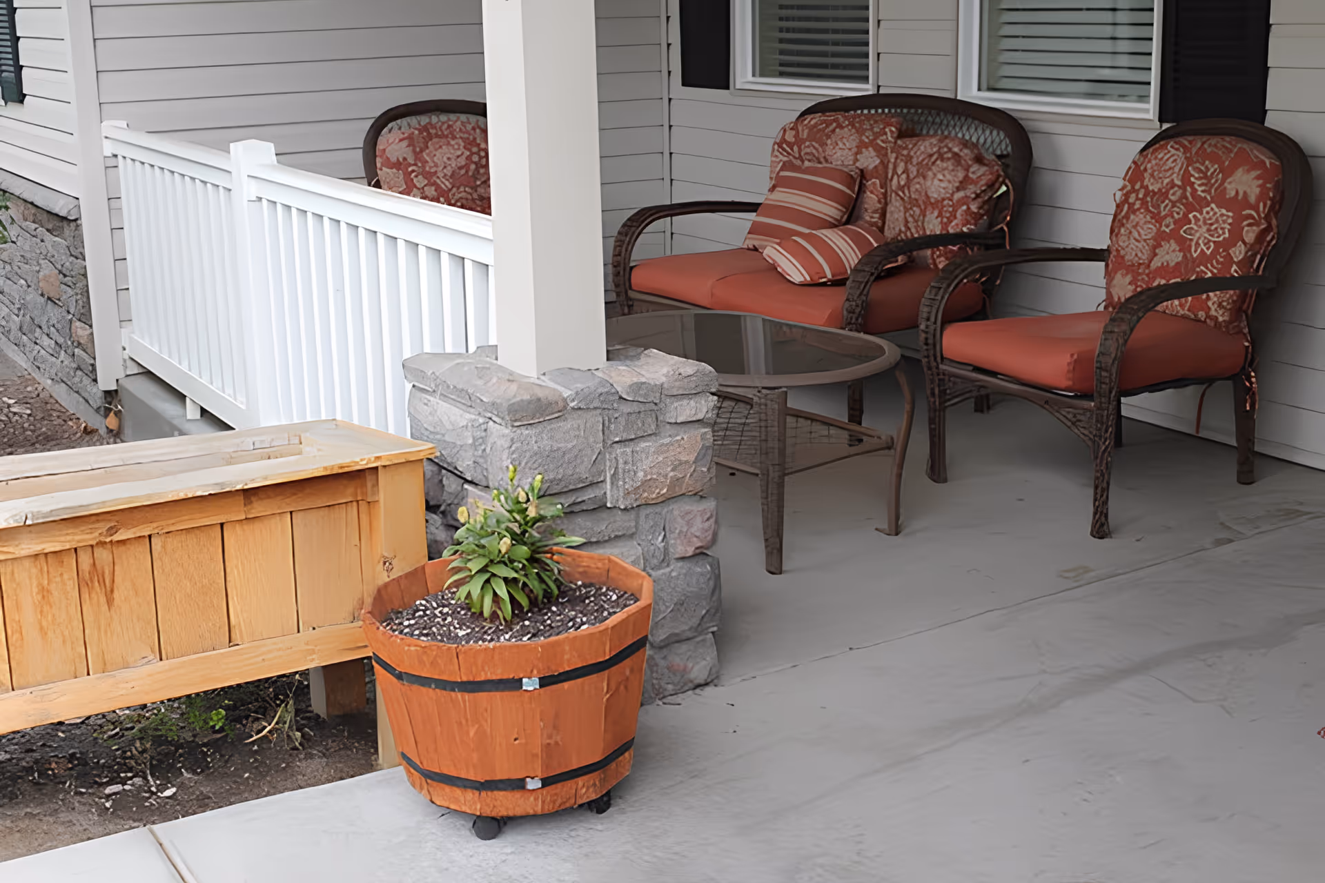 A covered outdoor patio area with two cushioned wicker chairs and a matching loveseat with floral and striped pillows. There is a glass-top coffee table between the seating. A wooden planter box and a round wooden pot with a small plant are in the foreground. The patio has a concrete floor and is adjacent to a white railing and stone pillar.