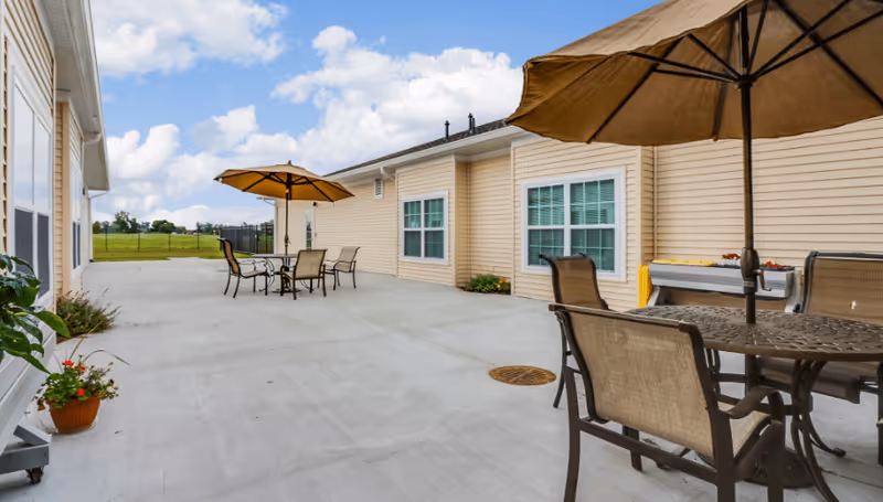 Outdoor patio area at Bayfield Assisted Living & Memory Care with beige siding buildings, concrete flooring, several tables with umbrellas, and chairs arranged for seating. There are potted plants near the building and a grassy field in the background under a partly cloudy sky.