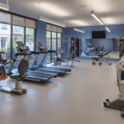 A modern fitness room with several treadmills, stationary bikes, weight machines, and exercise equipment. Large windows on one side let in natural light and offer a view of an outdoor patio area. A large mirror covers one wall, and a flat-screen TV is mounted above it.