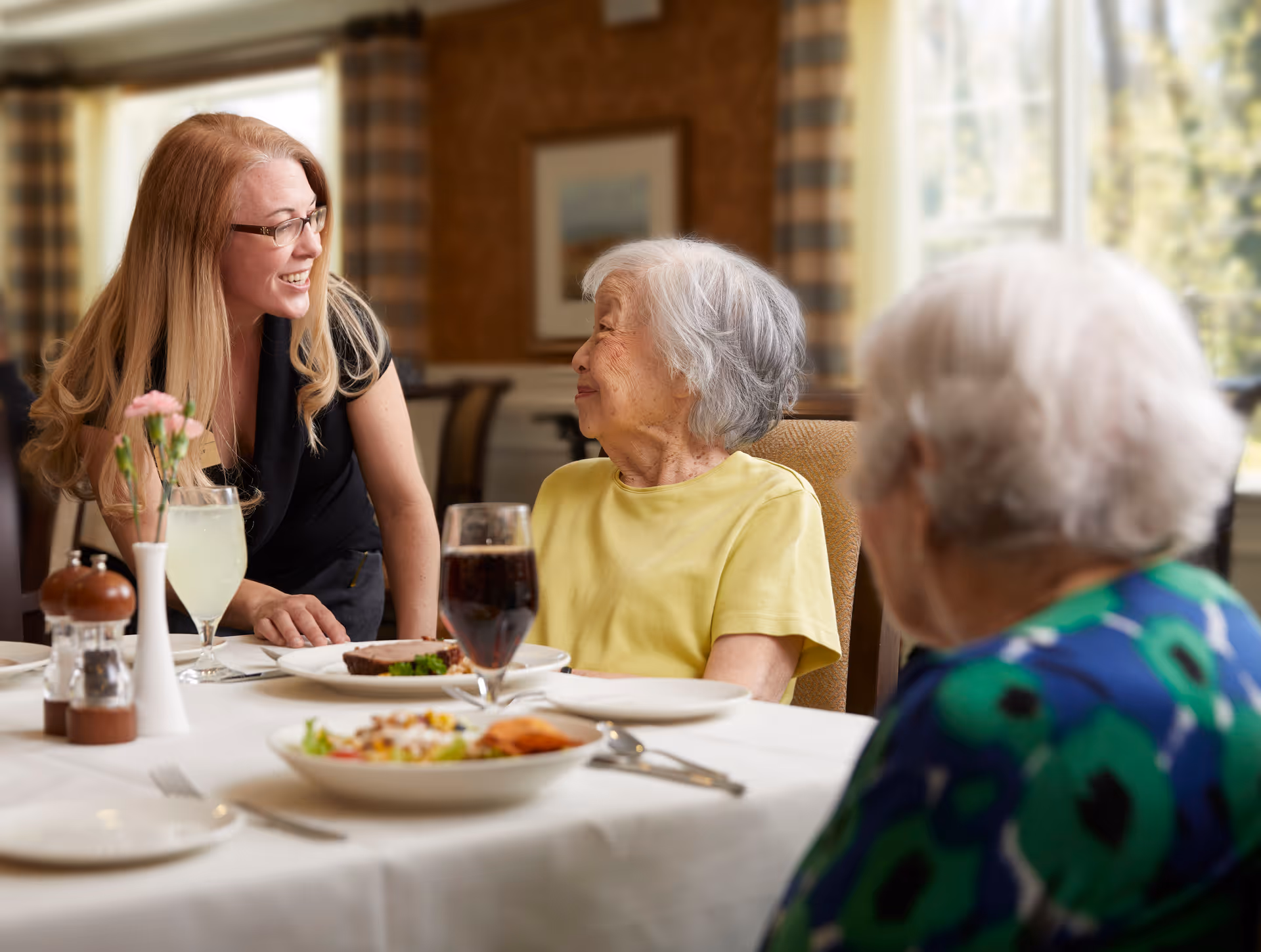 A smiling woman with long blonde hair and glasses leans toward two elderly women seated at a dining table in a warmly lit room. The elderly woman in a yellow shirt looks up at her, while the other elderly woman in a blue patterned top is partially visible. The table is set with plates of food, glasses of beverages, and a small vase with pink flowers.