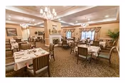 A spacious dining room with multiple tables covered in white tablecloths, each set with plates, cups, and napkins. The room features elegant chandeliers, a fireplace, large windows with drapes, and comfortable upholstered chairs. The decor is warm and inviting with beige walls and carpeted floors.