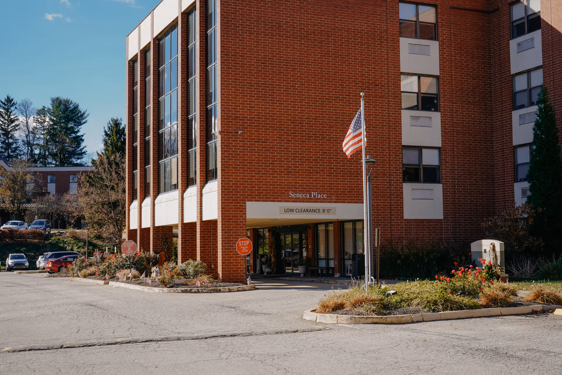 Exterior view of a brick senior living facility named Seneca Place with an American flag on a flagpole in front. The building has large windows and a covered entrance with a sign indicating low clearance of 8 feet. There are stop signs and landscaped areas with flowers and bushes near the entrance, and several parked cars are visible in the background.