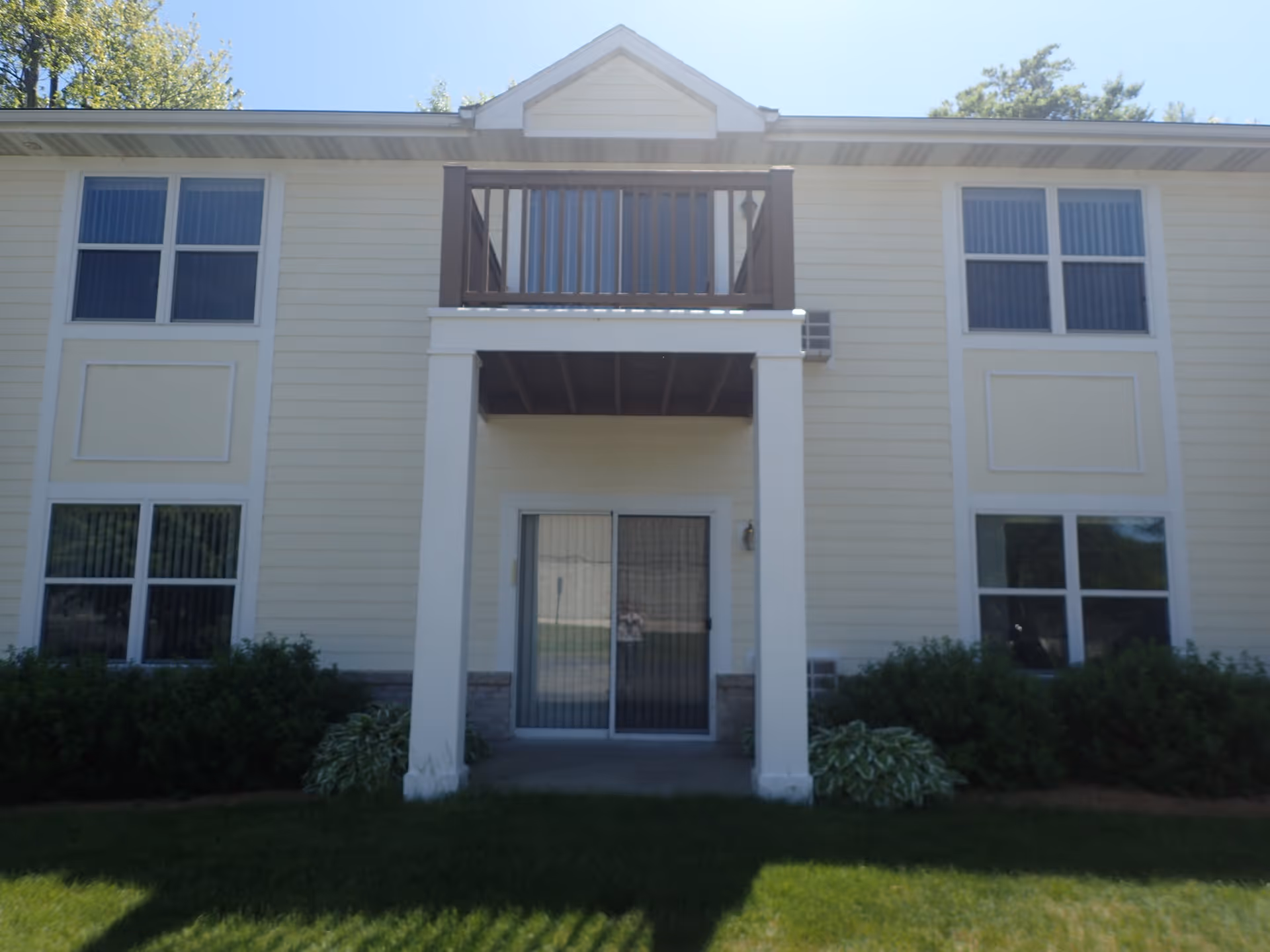 Exterior view of a two-story residential building with beige siding, white trim, and a small balcony above a covered patio area. The building has multiple windows with vertical blinds and is surrounded by green bushes and a well-maintained lawn under a clear blue sky.