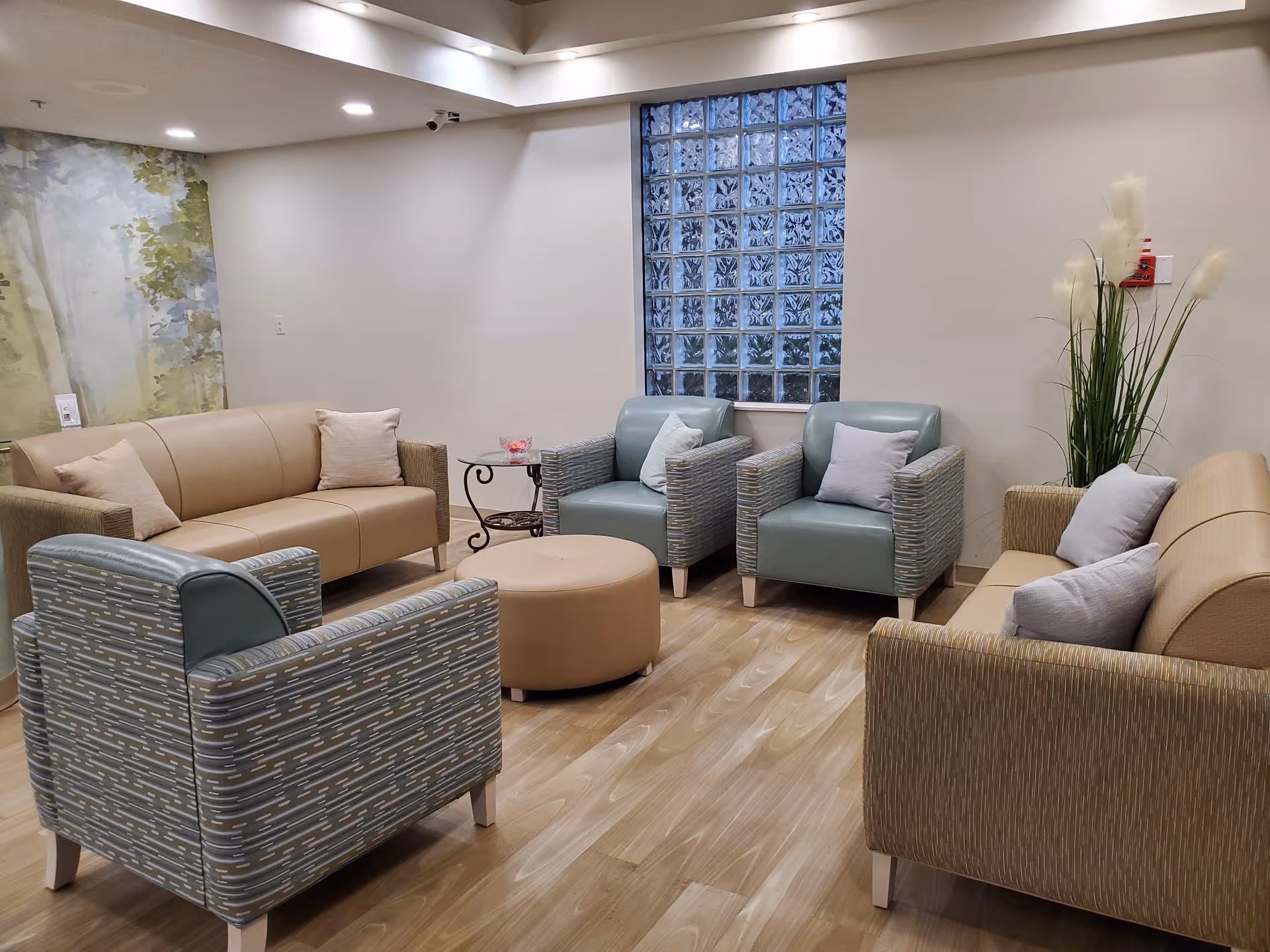 A cozy sitting area in a senior living facility with two beige sofas, two blue patterned armchairs, a round beige ottoman, a small glass side table, and decorative pillows. The room has light wood flooring, a wall with glass blocks, and a tall plant with white feathery flowers in the corner.