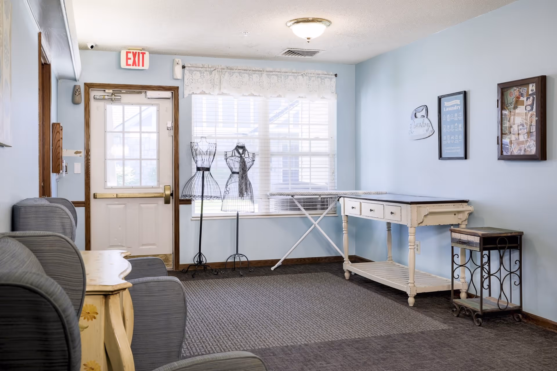 A light blue room with a white door featuring an exit sign above it. There are two wire dress forms near a window with white blinds and a lace valance. An ironing board is set up against the wall next to a white wooden table with drawers and a small decorative metal and wood cart. Three framed items hang on the wall, one with laundry instructions and another with various pinned notes. Several gray upholstered chairs are partially visible on the left side.