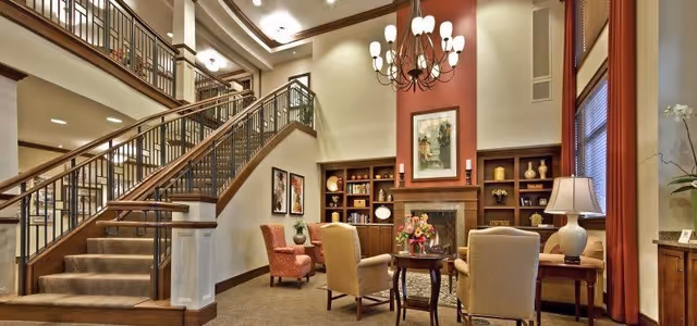 A spacious and warmly lit living room area in a senior living facility featuring a large staircase with wooden railings, comfortable armchairs arranged around a small round table with a flower vase, built-in wooden shelves with decorative items, a fireplace with a framed painting above it, and large windows with red curtains.