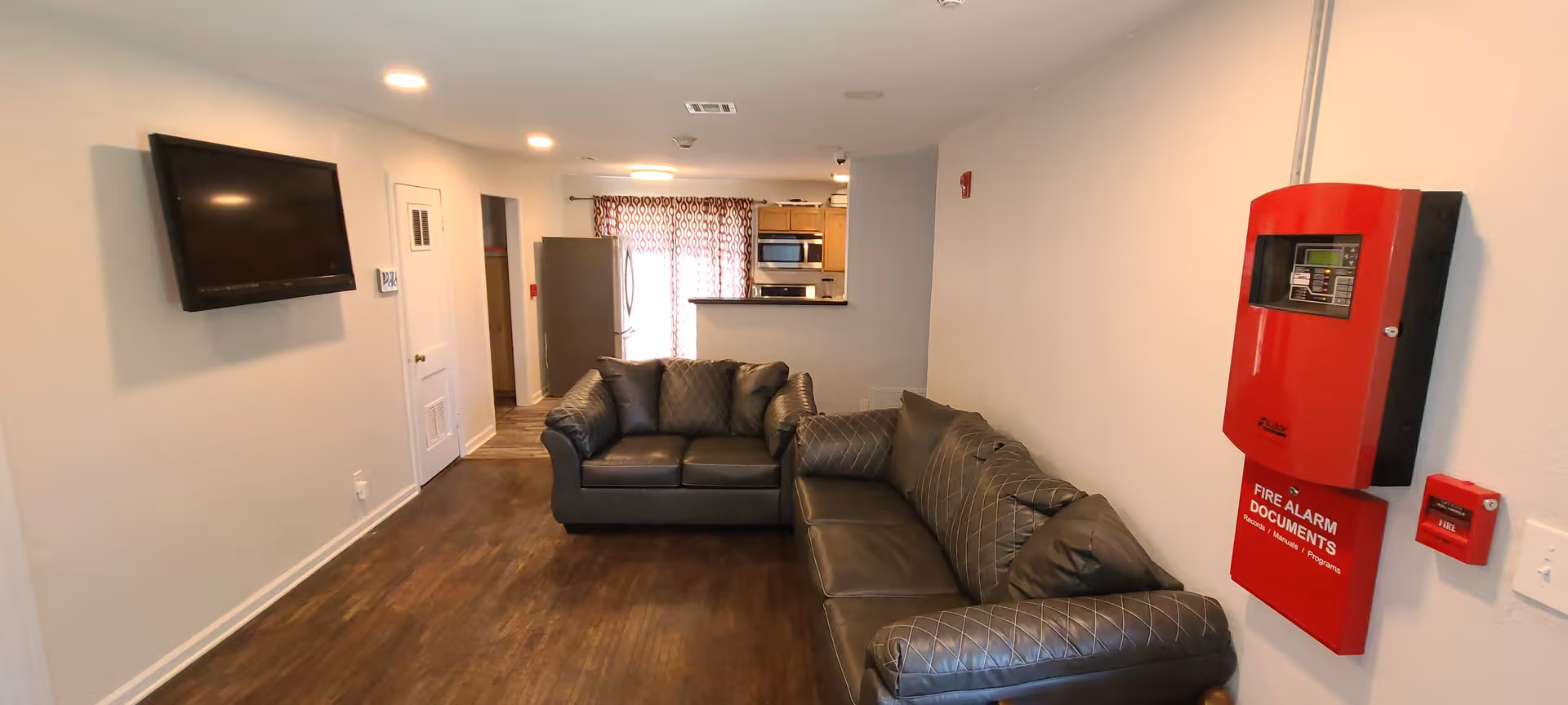 Long common living area with leather sofas, a wall-mounted TV, a view into the kitchen, and a red fire-alarm box on the wall.