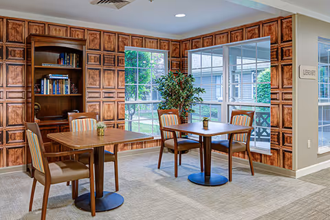 A cozy interior room with two wooden tables, each surrounded by four chairs with striped upholstery. The walls feature a decorative wood panel design with built-in shelves holding books. Large windows let in natural light and show greenery outside. A potted plant is placed near the windows, and a sign on the wall reads 'Library'.