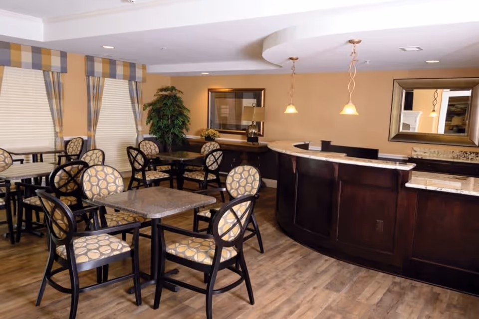 A dining area with multiple tables and chairs featuring patterned upholstery. The room has wood flooring, beige walls, and windows with plaid valances. There is a curved dark wood reception or service counter with a marble countertop, two pendant lights hanging above it, and a large mirror on the wall behind the counter. A potted plant and a table lamp are also visible in the background.