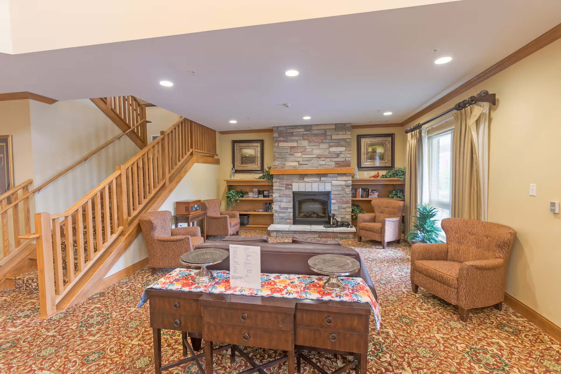 A cozy living room area with a stone fireplace centered on the far wall, flanked by built-in wooden shelves with plants and framed artwork above. Four upholstered armchairs are arranged around the fireplace, with a wooden table covered by a colorful floral runner in the foreground. A wooden staircase with railings is visible on the left side, and a window with beige curtains is on the right wall. The room has warm yellow walls and a patterned carpet.