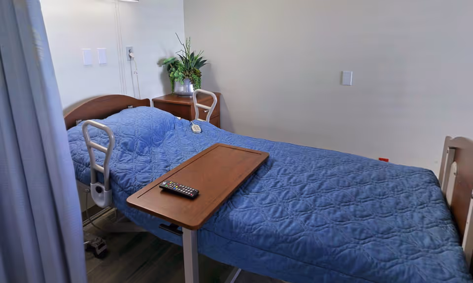 A hospital-style bed with a blue quilted blanket in a room. The bed has side rails and an overbed table with a TV remote on it. There is a small wooden nightstand with a plant in a glass vase next to the bed. The room has plain white walls and a curtain partially visible on the left side.