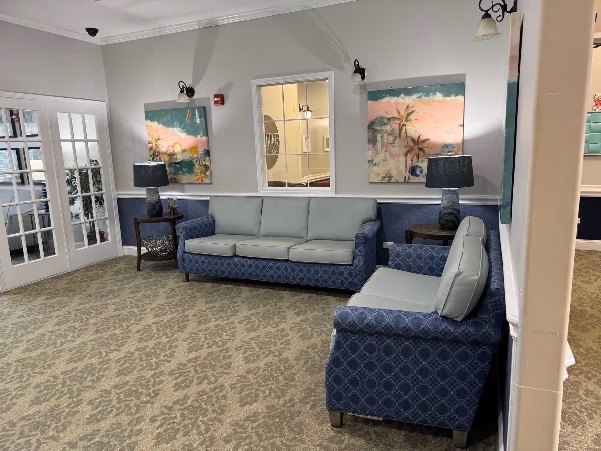 A cozy sitting area in a senior living facility with two blue patterned sofas with light gray cushions arranged around a corner. There are two side tables with matching blue lamps and colorful artwork featuring palm trees on the walls. A window looks into another room, and French doors are visible on the left side. The carpet has a floral pattern in beige tones.