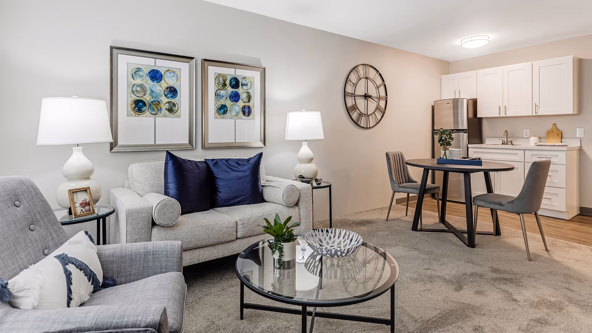 A modern living room and kitchenette area featuring a light gray sofa with two dark blue pillows, a gray armchair with a white and blue pillow, a round glass coffee table with decorative items, two table lamps on side tables, two framed abstract artworks on the wall, a round dining table with two gray chairs, and a kitchenette with white cabinets and stainless steel refrigerator.
