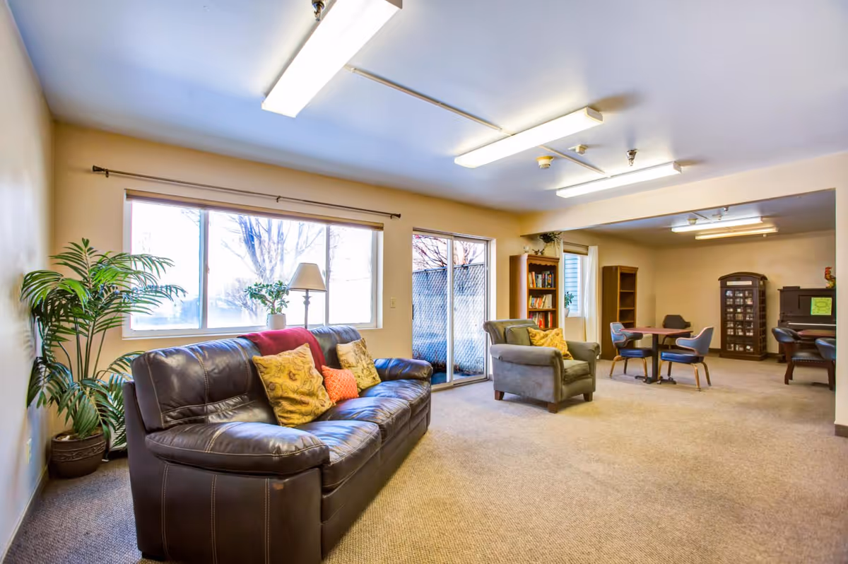 A bright senior living common area with a dark leather sofa adorned with colorful pillows, a gray armchair, a potted plant, a floor lamp, large windows, and a sliding glass door leading outside. In the background, there are tables with chairs, bookshelves, and additional seating.