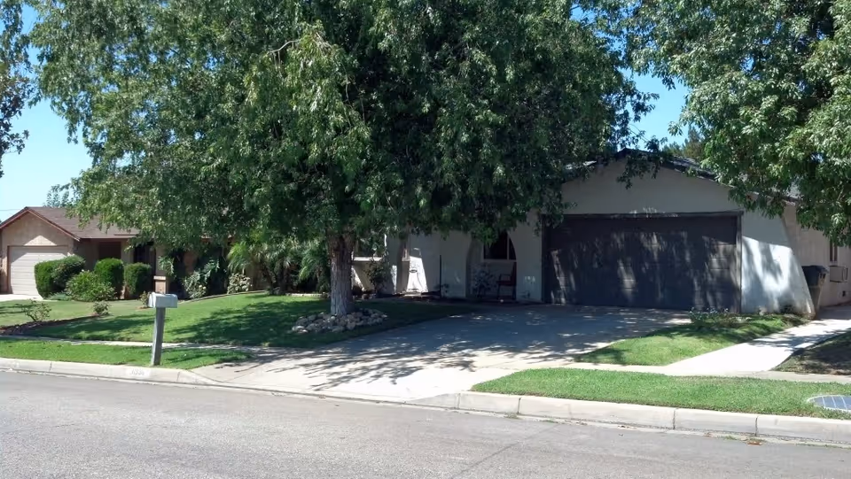 Street view of a single-story house with a large tree in the front yard and a driveway leading to a two-car garage.
