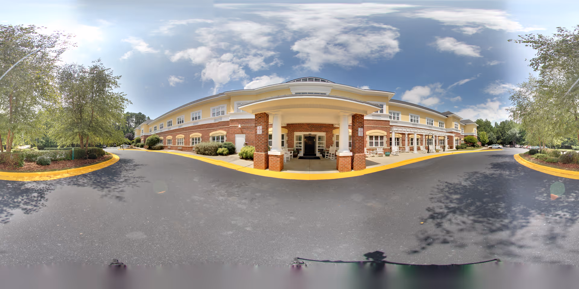Wide panoramic view of the front exterior of Fox Hollow Senior Living Community building with a covered entrance supported by brick columns, surrounded by trees and landscaping under a partly cloudy sky.