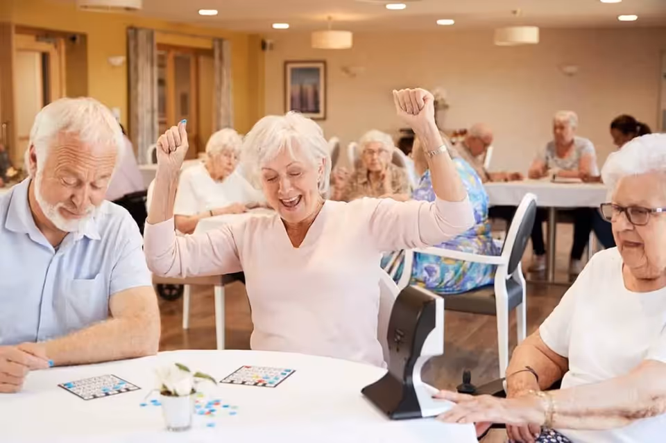 A group of elderly people sitting around tables in a well-lit room, playing bingo. One woman in the center is raising her arms in celebration, appearing happy and excited. Other seniors are focused on their bingo cards.