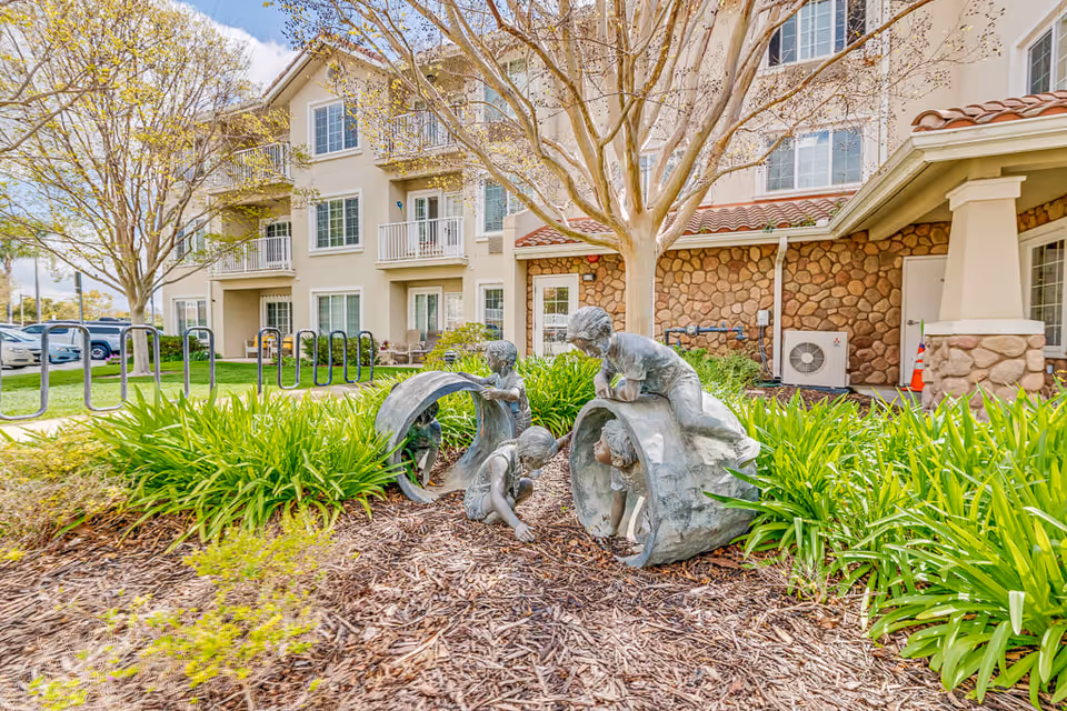 Outdoor garden area in front of a multi-story assisted living building with stone and beige exterior walls. The garden features a sculpture of children playing inside and around large hollow cylindrical shapes, surrounded by green plants and mulch. Trees with budding leaves are visible, along with parked cars and bike racks in the background.