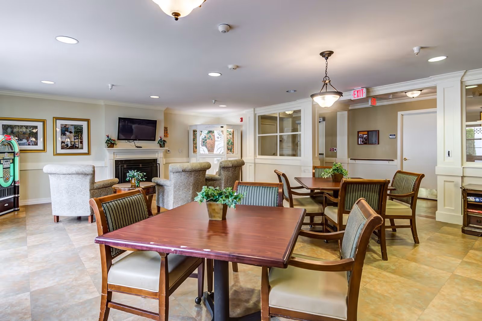 A well-lit common area with wooden tables and cushioned chairs arranged for seating. In the background, there are comfortable armchairs facing a wall-mounted television above a fireplace. The room has framed pictures on the walls, a jukebox on the left side, and potted plants on the tables. The ceiling has recessed lighting and a hanging light fixture, with an exit sign visible near a doorway.