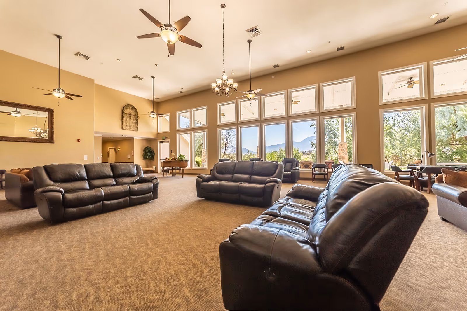 Spacious senior living facility common area with multiple black leather sofas arranged on carpeted floor, large windows letting in natural light and offering a view of trees and mountains, ceiling fans and chandeliers hanging from the high ceiling, and additional seating and tables near the windows.