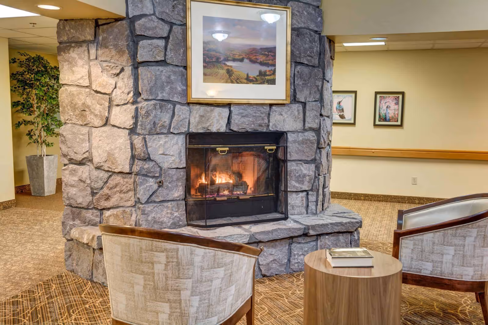 Cozy seating area in a senior living facility featuring a stone fireplace with a fire burning inside. Two upholstered chairs with wooden frames face the fireplace, with a small round wooden table between them holding a book. The walls are painted beige with framed artwork and a potted plant in the background.