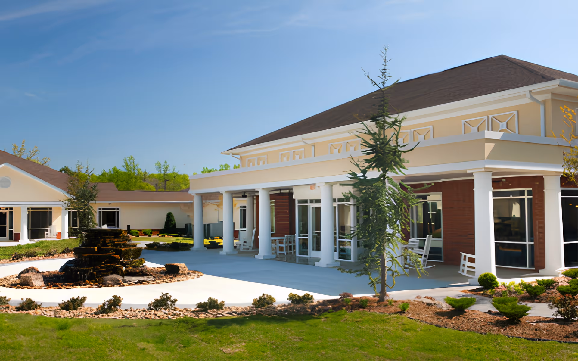 Front exterior of a senior living facility with a covered columned entry, rocking chairs, and a landscaped courtyard with a fountain.