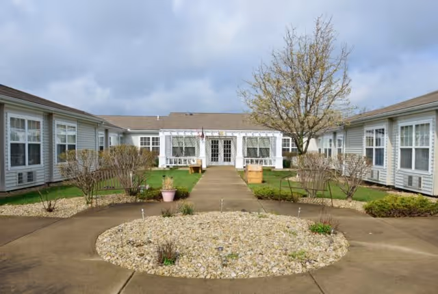Outdoor courtyard area of a senior living facility with a circular rock garden in the center, surrounded by concrete walkways, small bushes, and a tree. The building has white siding and multiple windows, with a covered entrance in the background under a cloudy sky.