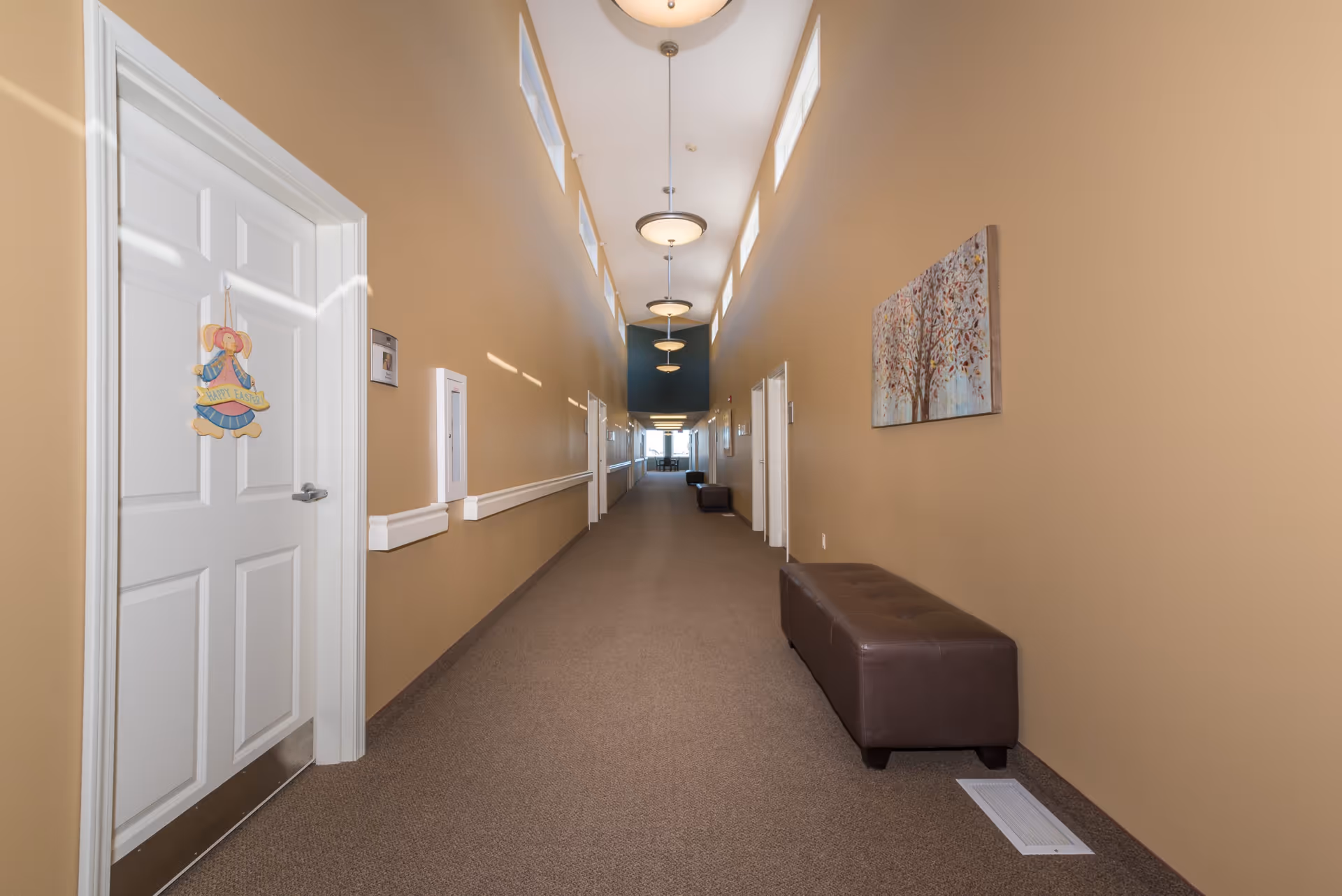 A long, well-lit hallway in a senior living facility with beige walls and carpeted floor. The hallway has multiple white doors on both sides, one decorated with a hanging sign that says 'Happy Easter' featuring a bunny. There are brown cushioned benches along the right wall and a painting of a tree. Ceiling lights and small windows near the ceiling provide illumination.