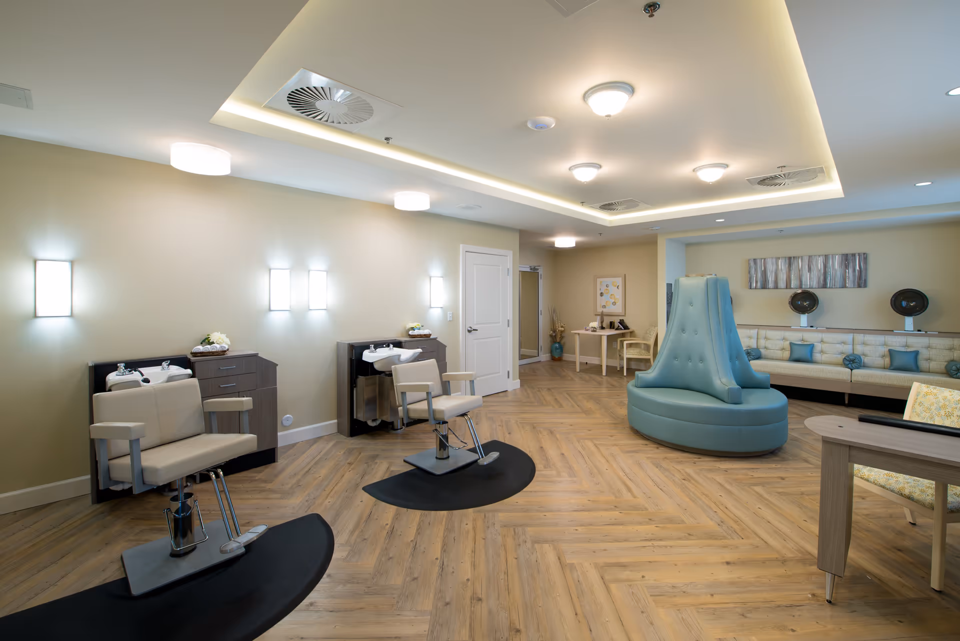 A spacious and well-lit salon area in a senior living facility featuring two salon chairs with sinks for hair washing, a large teal cushioned seating area in the center, a long beige couch with blue pillows along the back wall, and a small table with chairs in the background. The room has wood-patterned flooring and soft yellow walls with modern ceiling lights.