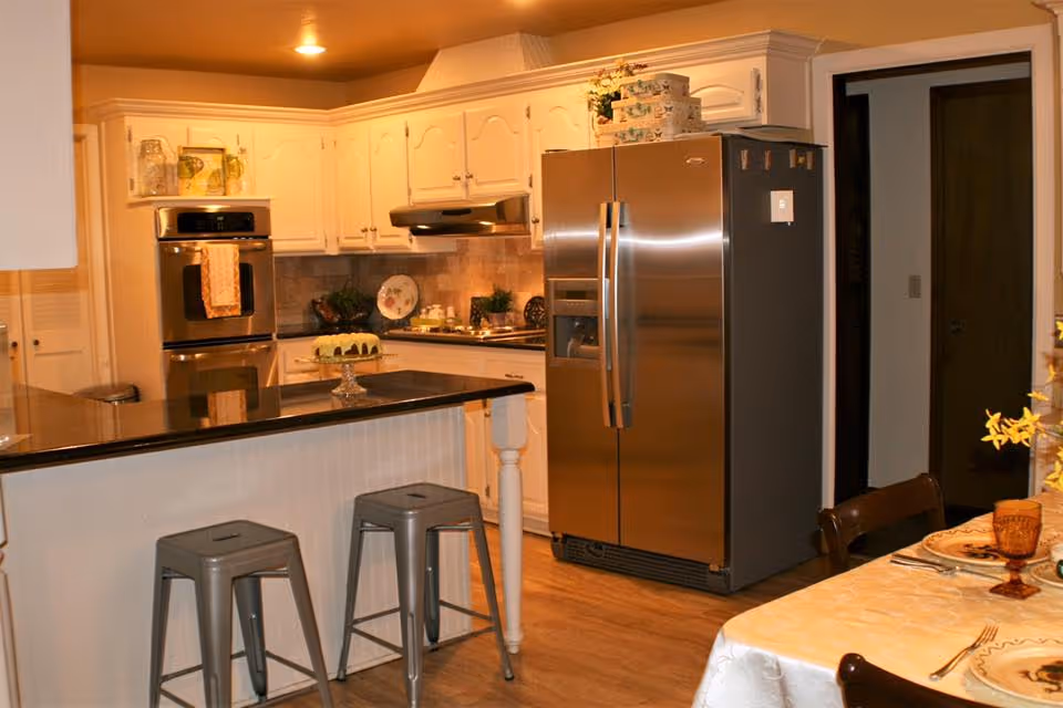 A warm, well-lit kitchen featuring white cabinetry, a stainless steel double-door refrigerator, a built-in oven, and a black countertop island with two metal stools. A cake is displayed on a glass cake stand on the island. To the right, a dining table is partially visible with a white tablecloth, plates, cutlery, and amber-colored glasses.