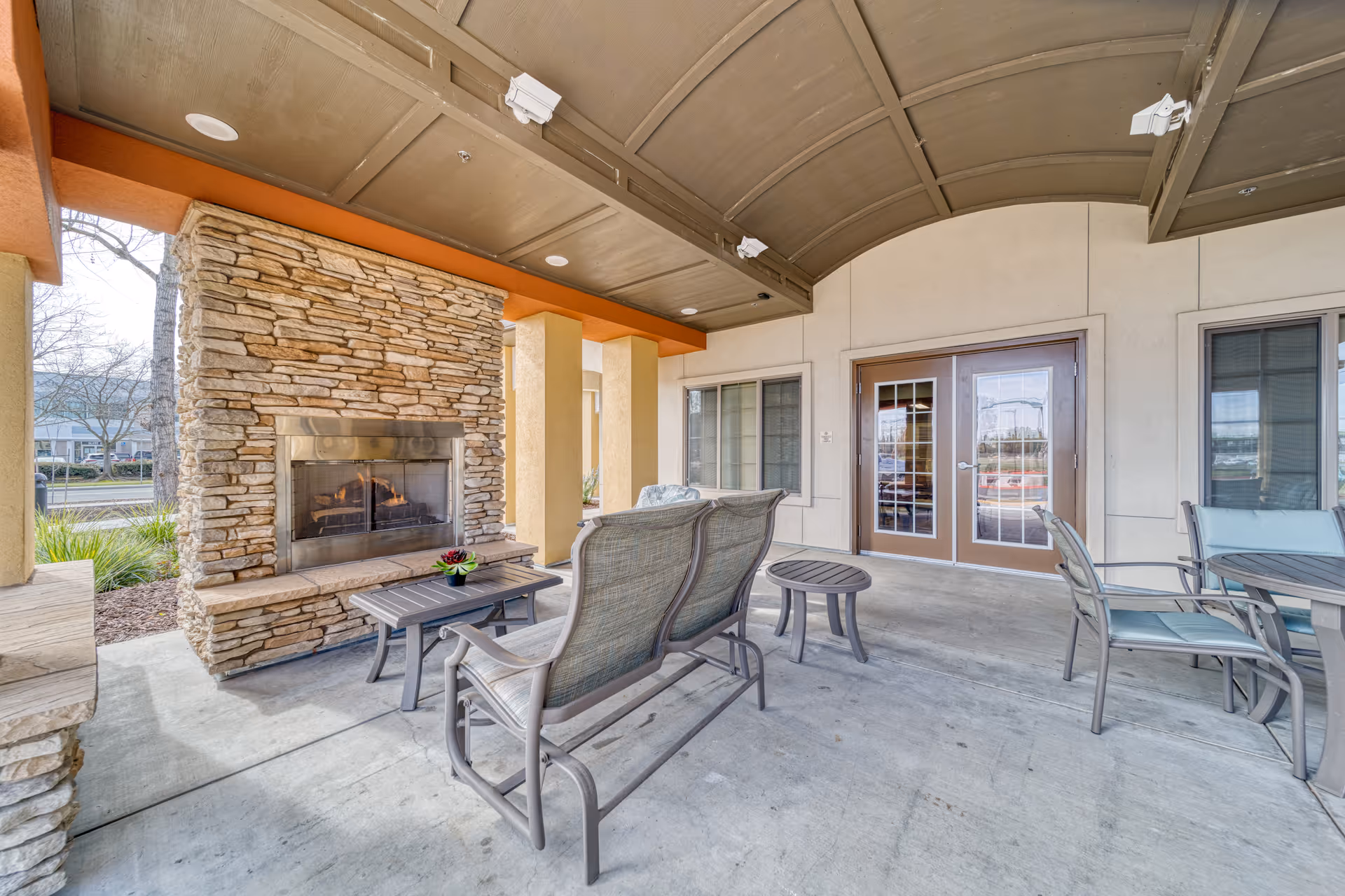 Covered outdoor seating area with a stone fireplace, cushioned chairs, a small table with a plant, and double glass doors leading inside the building.