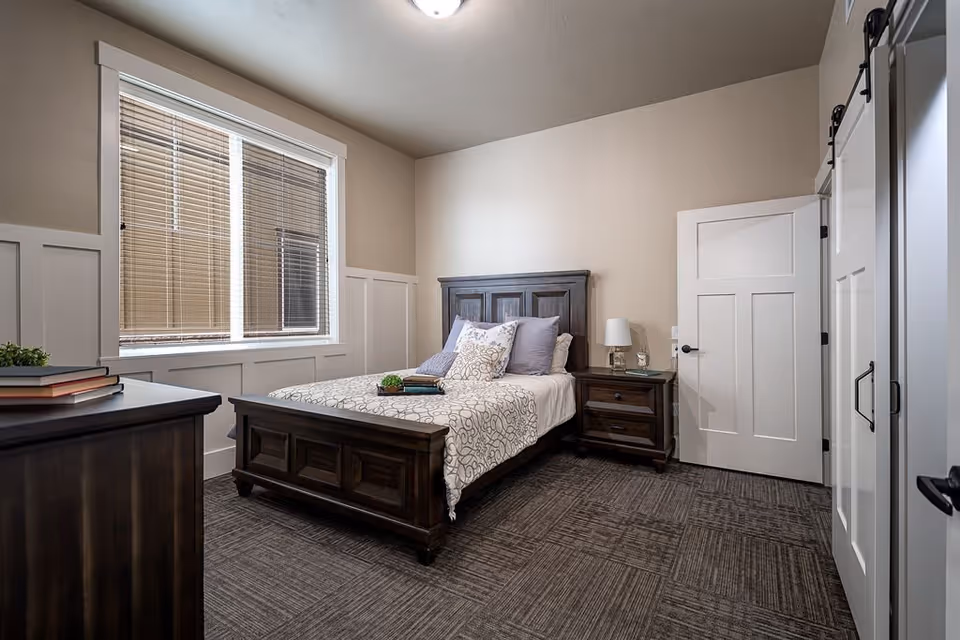 A neatly arranged bedroom in a senior living facility featuring a dark wooden bed with patterned bedding and multiple pillows. There is a matching nightstand with a lamp and a small decorative item beside the bed. A large window with blinds allows natural light into the room. The room has beige walls, a carpeted floor, and white doors, one of which is a sliding barn-style door.