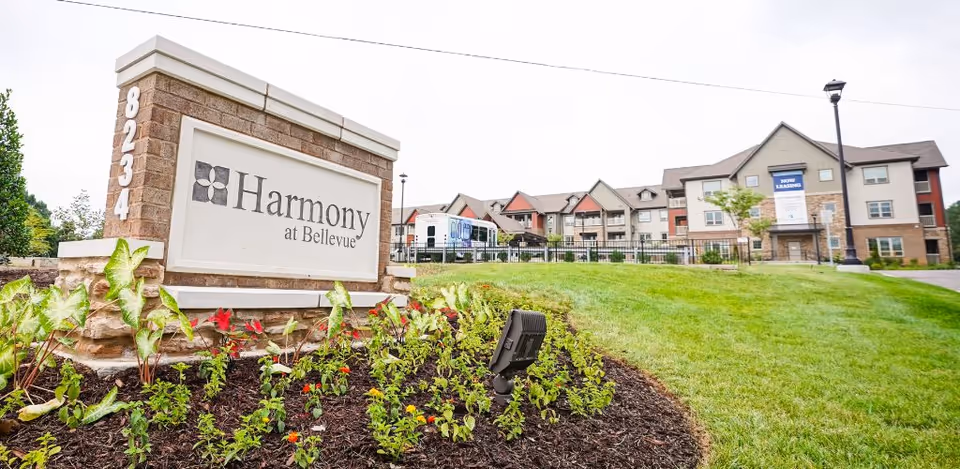 Stone entrance sign reading "Harmony at Bellevue" set in a landscaped bed with the multi-story facility building and lawn behind it.