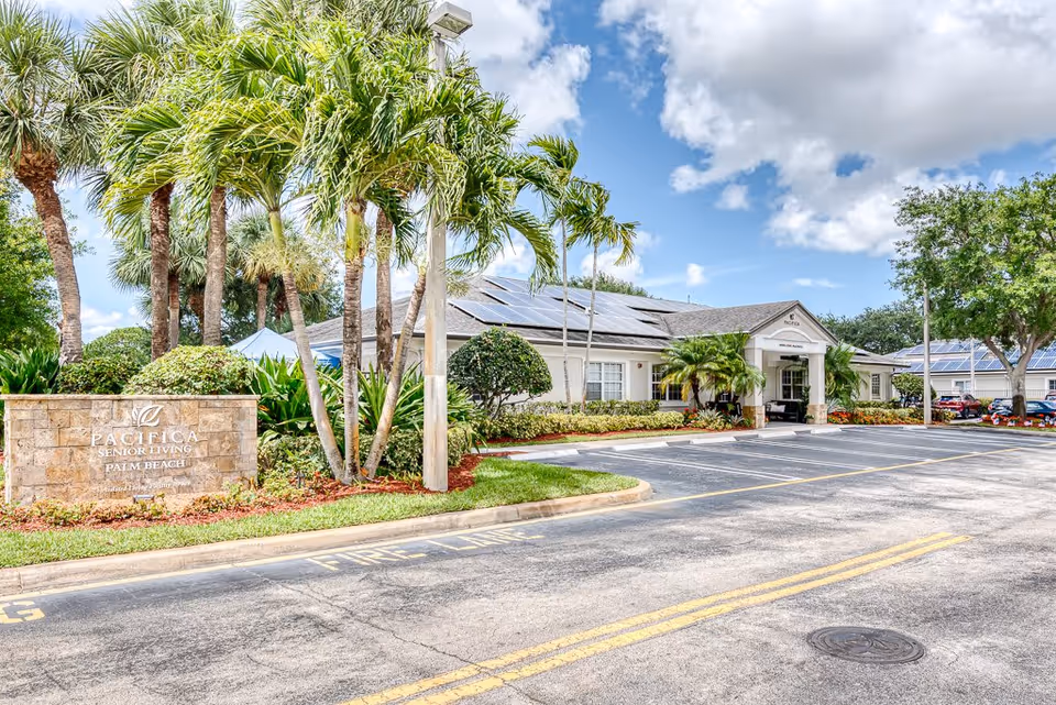 Exterior view of Pacifica Senior Living Palm Beach facility with a stone sign, palm trees, landscaped bushes, and a parking lot under a partly cloudy sky.
