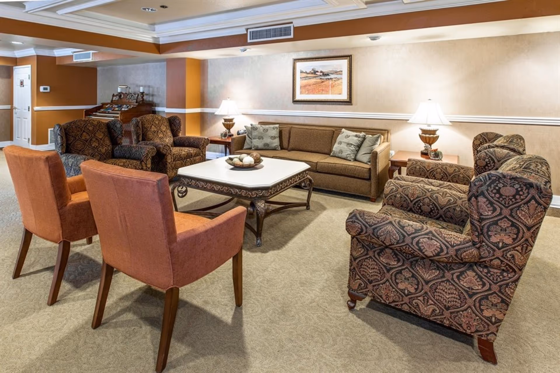 A cozy living room area with a beige sofa adorned with patterned pillows, two orange upholstered chairs, and three patterned armchairs arranged around a square coffee table with decorative items. The room features beige walls with a white chair rail, a framed landscape painting, two table lamps on side tables, and a carpeted floor.