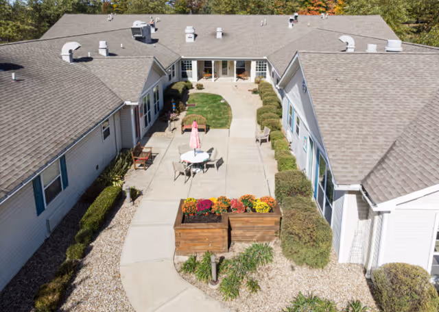 Aerial view of a courtyard area at Arden Courts - ProMedica Memory Care Community (Parma) featuring a paved walkway, outdoor seating with a table and umbrella, wooden planters with colorful flowers, and surrounding single-story buildings with gray shingled roofs and white siding.