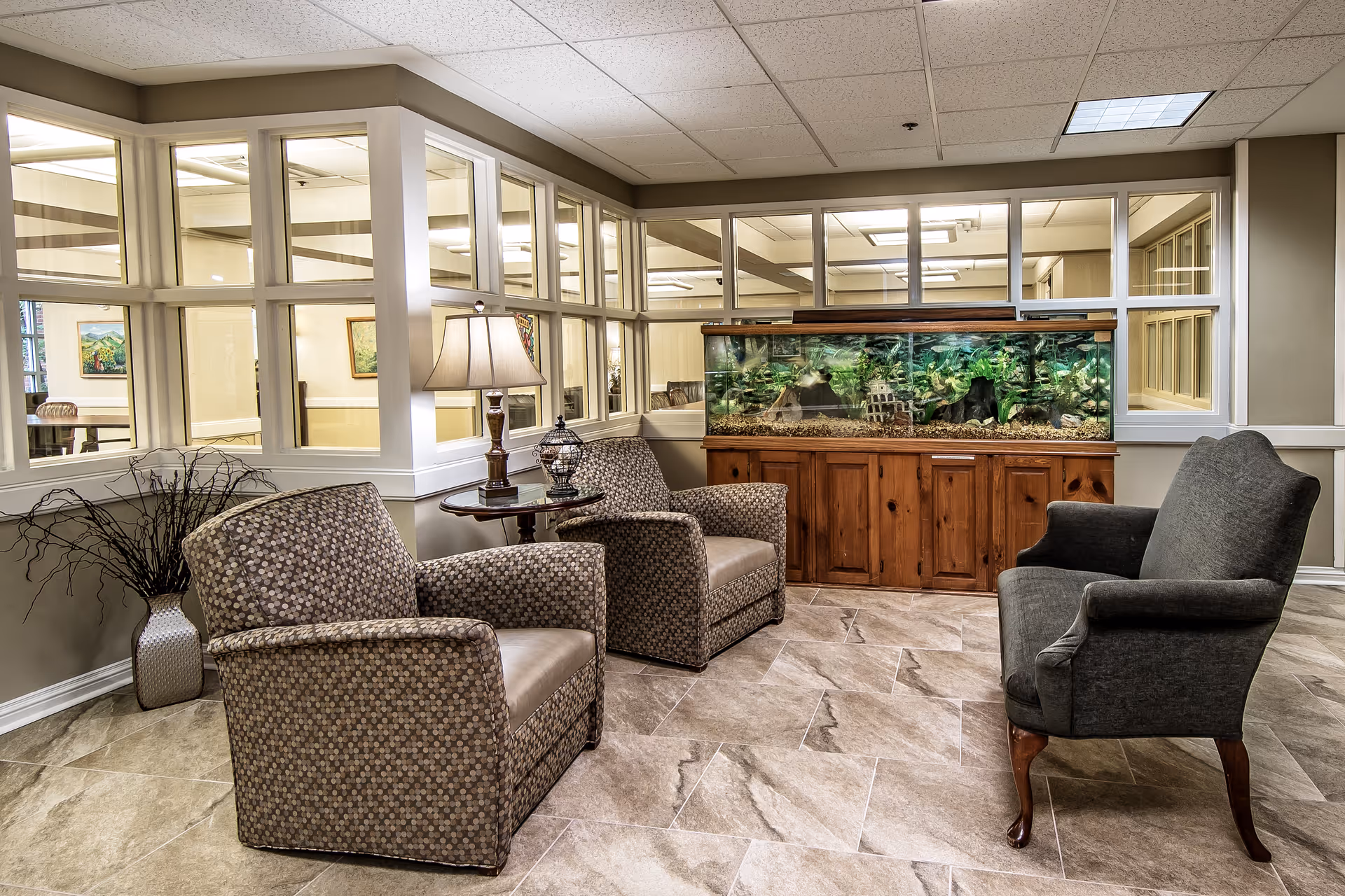 A cozy sitting area in a senior living facility with three upholstered armchairs arranged around a wooden side table with a lamp. Behind the chairs is a large fish tank set on a wooden cabinet. The room has beige tiled flooring and walls with large windows looking into adjacent rooms.