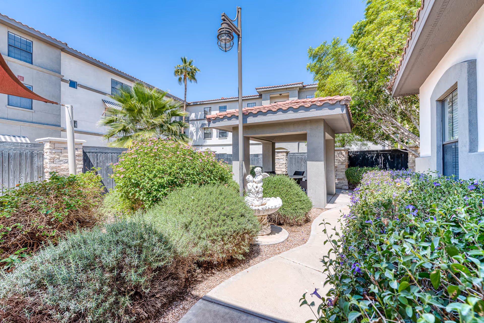 Outdoor garden area at The Woodmark at Sun City featuring a paved walkway, lush green bushes, flowering plants, a small decorative fountain with cherub statues, a covered seating area with a tiled roof, and a tall street lamp under a clear blue sky.