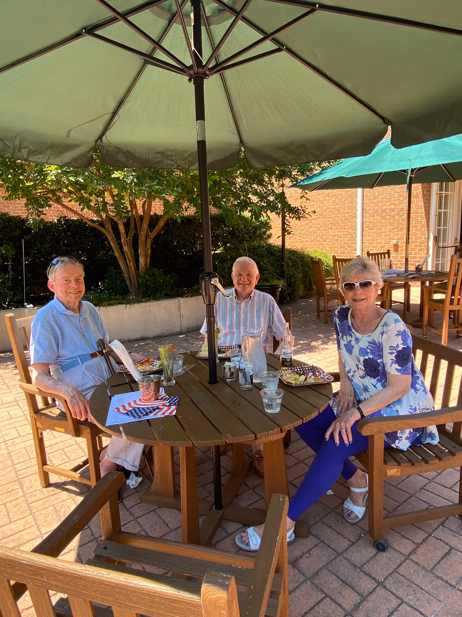 Three elderly people sitting around a wooden outdoor table with a large green umbrella providing shade. They are enjoying a meal and drinks on a sunny day in a patio area with brick flooring and greenery in the background.